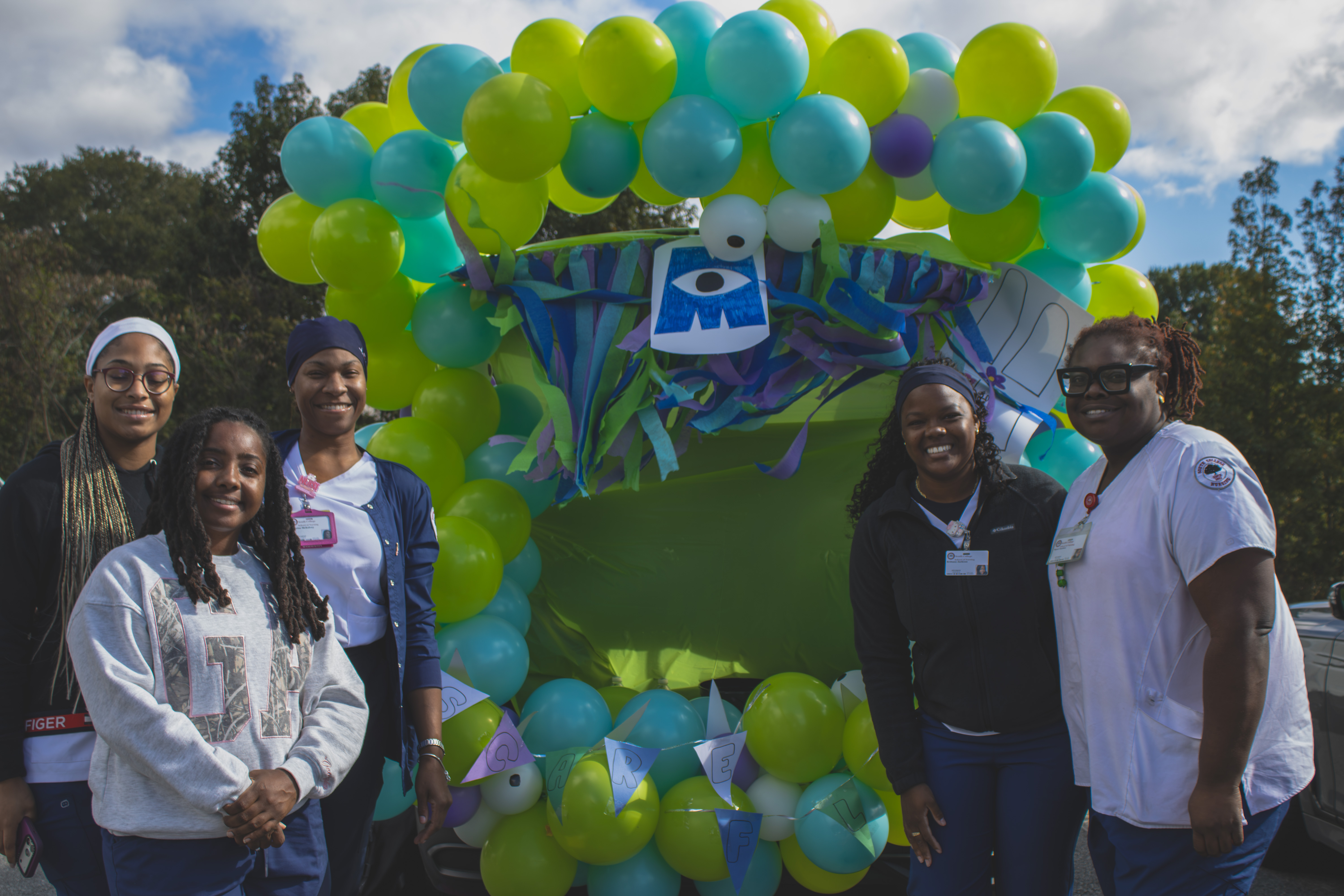 Five people pose beside a Monsters, Inc.–themed trunk decorated with green and turquoise balloons.